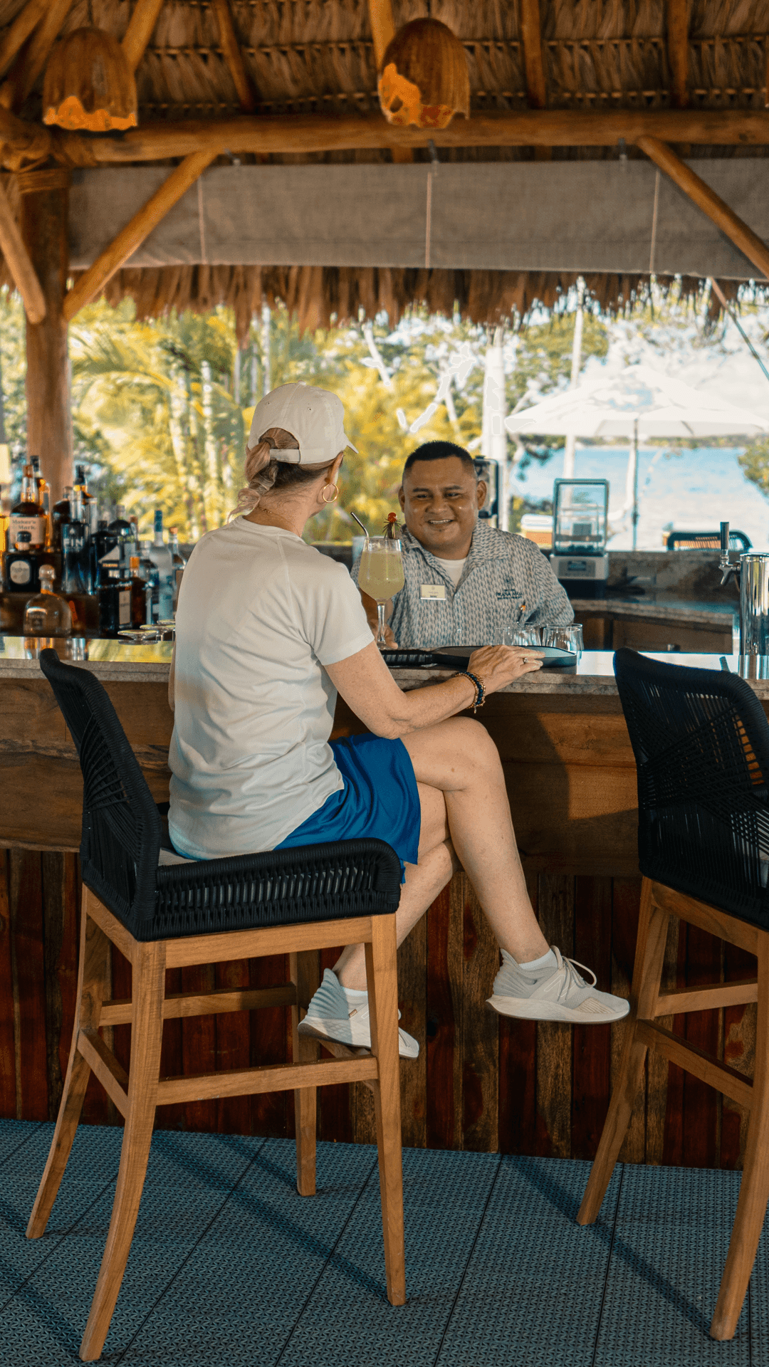 A person in a white cap sits at a bar chatting with a smiling bartender.