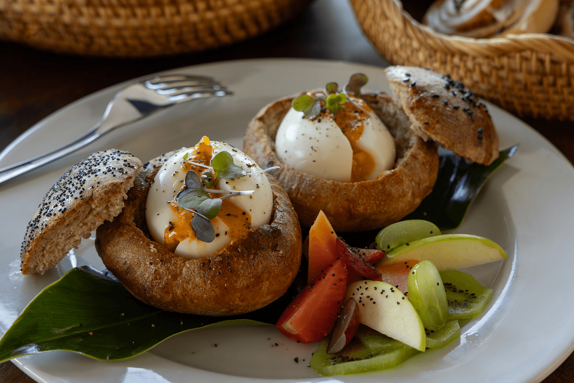 A plate featuring two baked bread bowls filled with poached eggs, garnished with microgreens, alongside a colorful fruit arrangement.