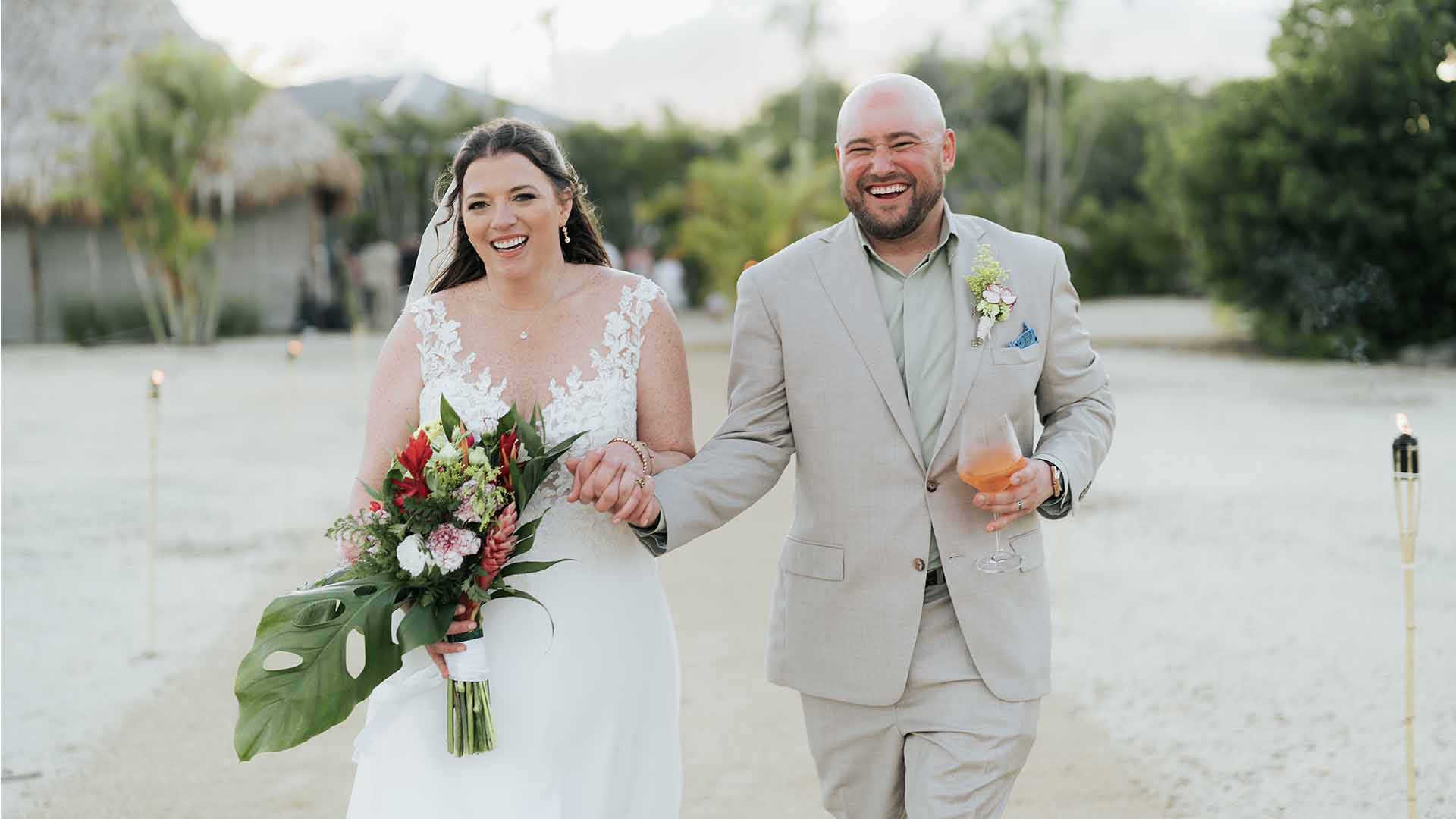 A bride and groom joyfully walk hand-in-hand along a sandy path, smiling and holding a bouquet and drink.