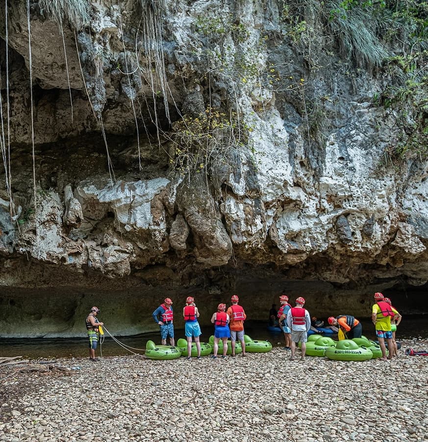 A group of people in life jackets gathers near kayaks under a rocky overhang by a riverbank.