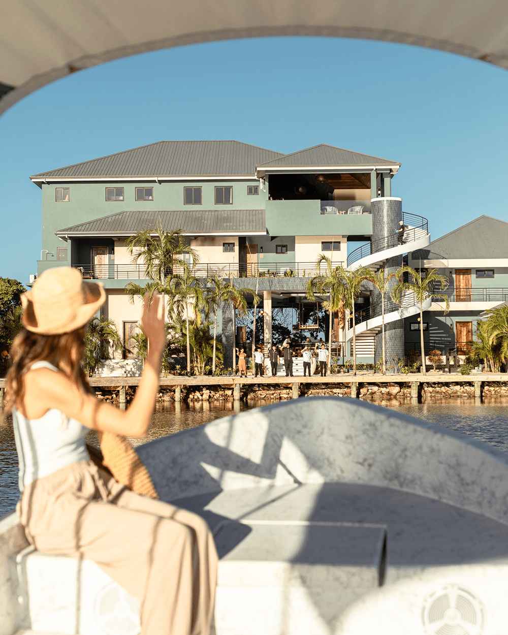 A person in a boat waves towards a modern waterfront building surrounded by palm trees.