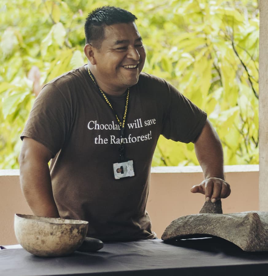 A smiling man in a brown shirt promoting chocolate's role in saving the rainforest, standing by traditional tools.