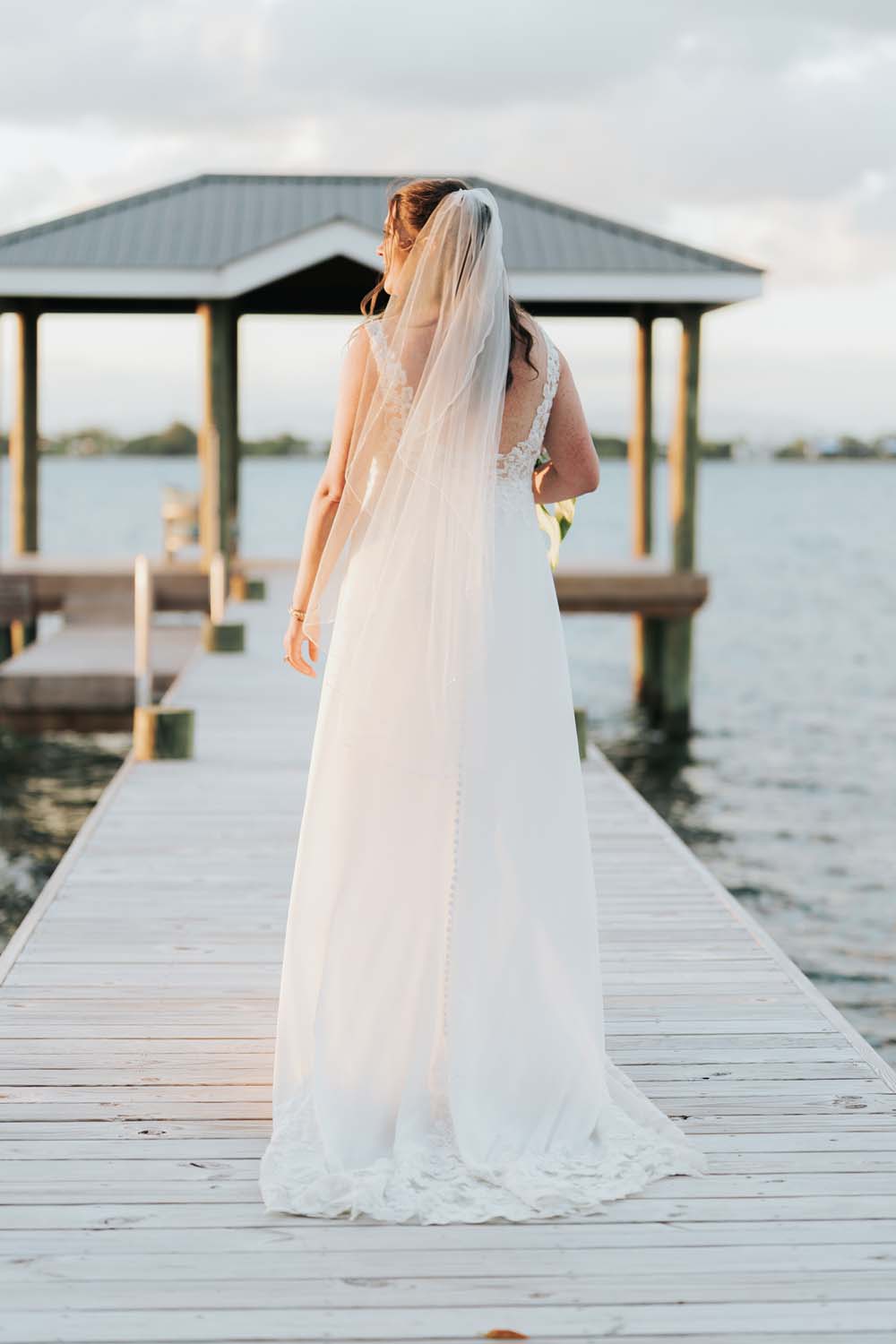A bride in a white gown walks down a wooden dock towards the water.