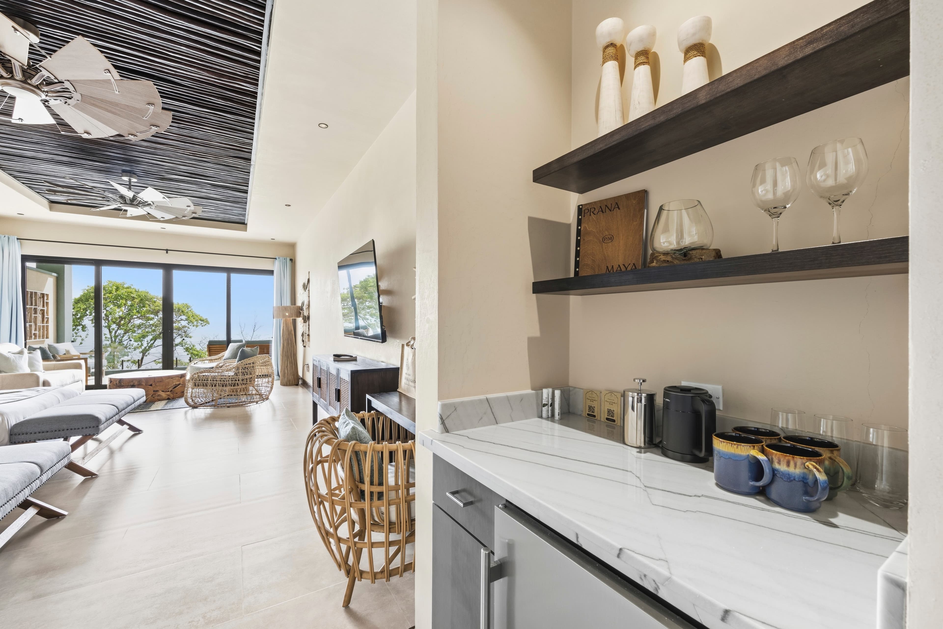 A modern, airy living space features a kitchenette with a marble counter and a view of greenery through large windows.