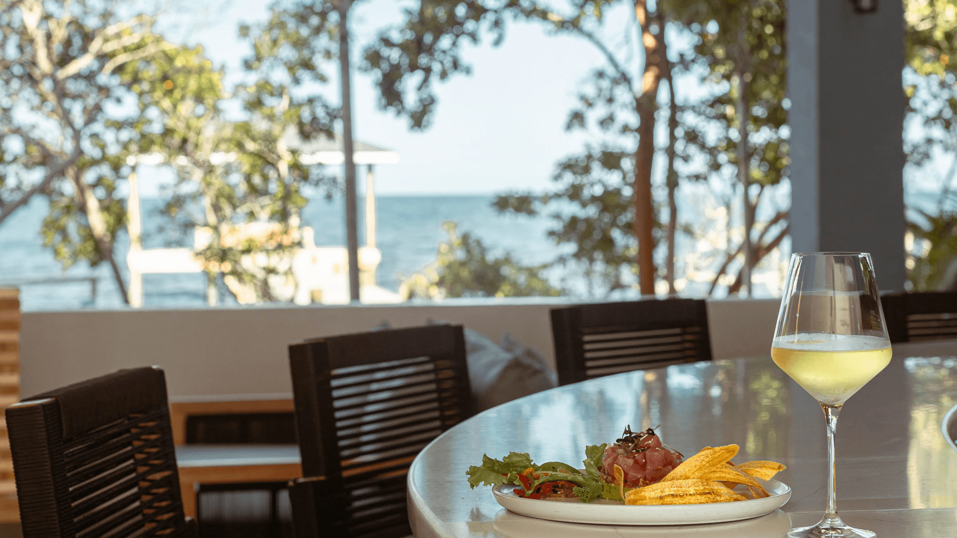 A stylish plate of food with plantains and salad sits on a table beside a glass of drink, overlooking a scenic ocean view.