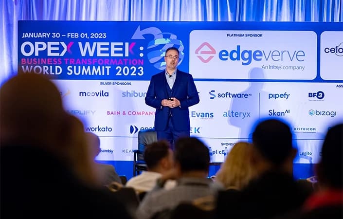 A speaker stands on stage at the OPEX Week: Business Transformation World Summit 2023, with a backdrop of logos from various sponsoring companies.