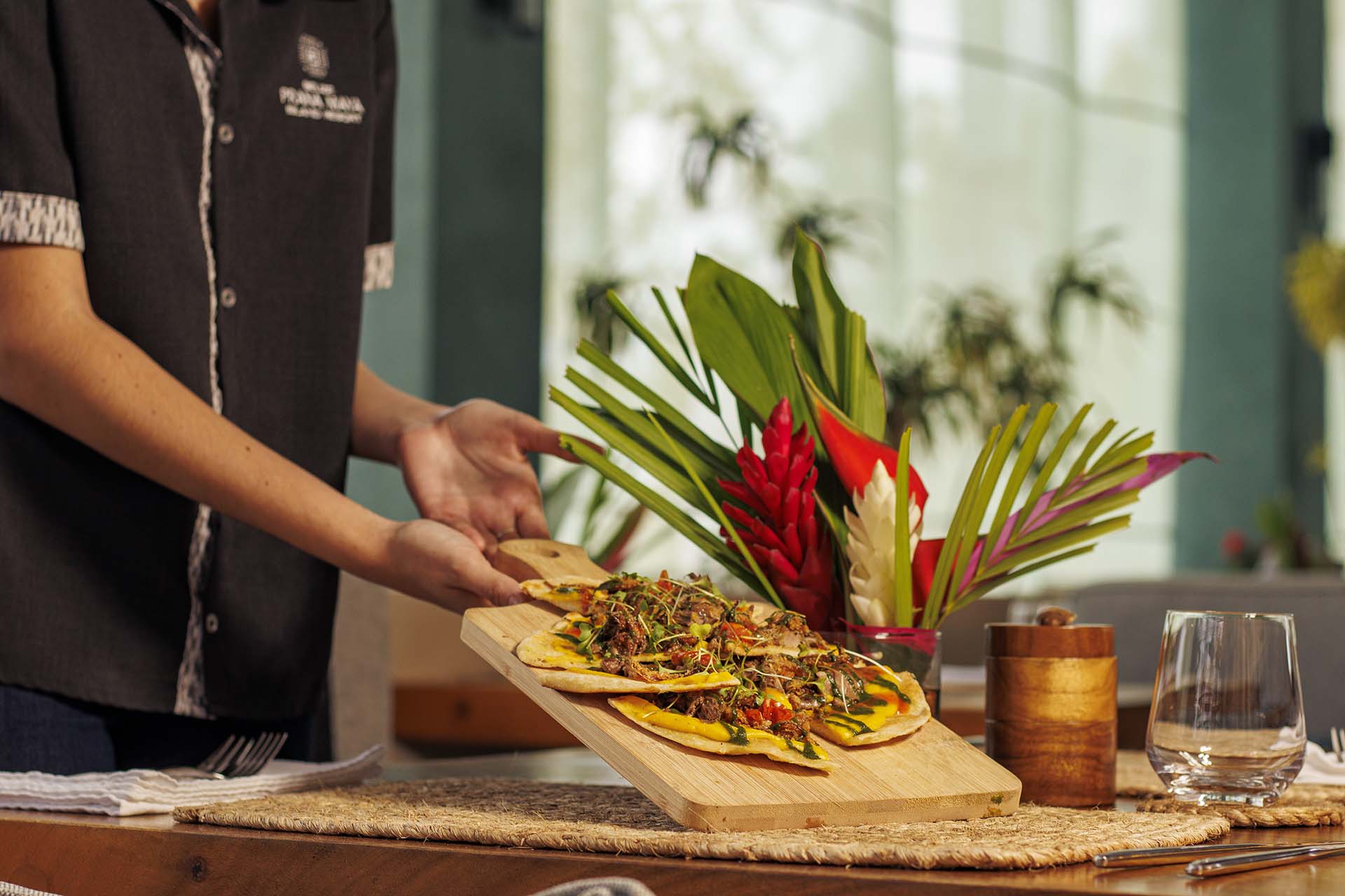 A server presents a wooden platter with a colorful dish, surrounded by tropical plants in a dining setting.