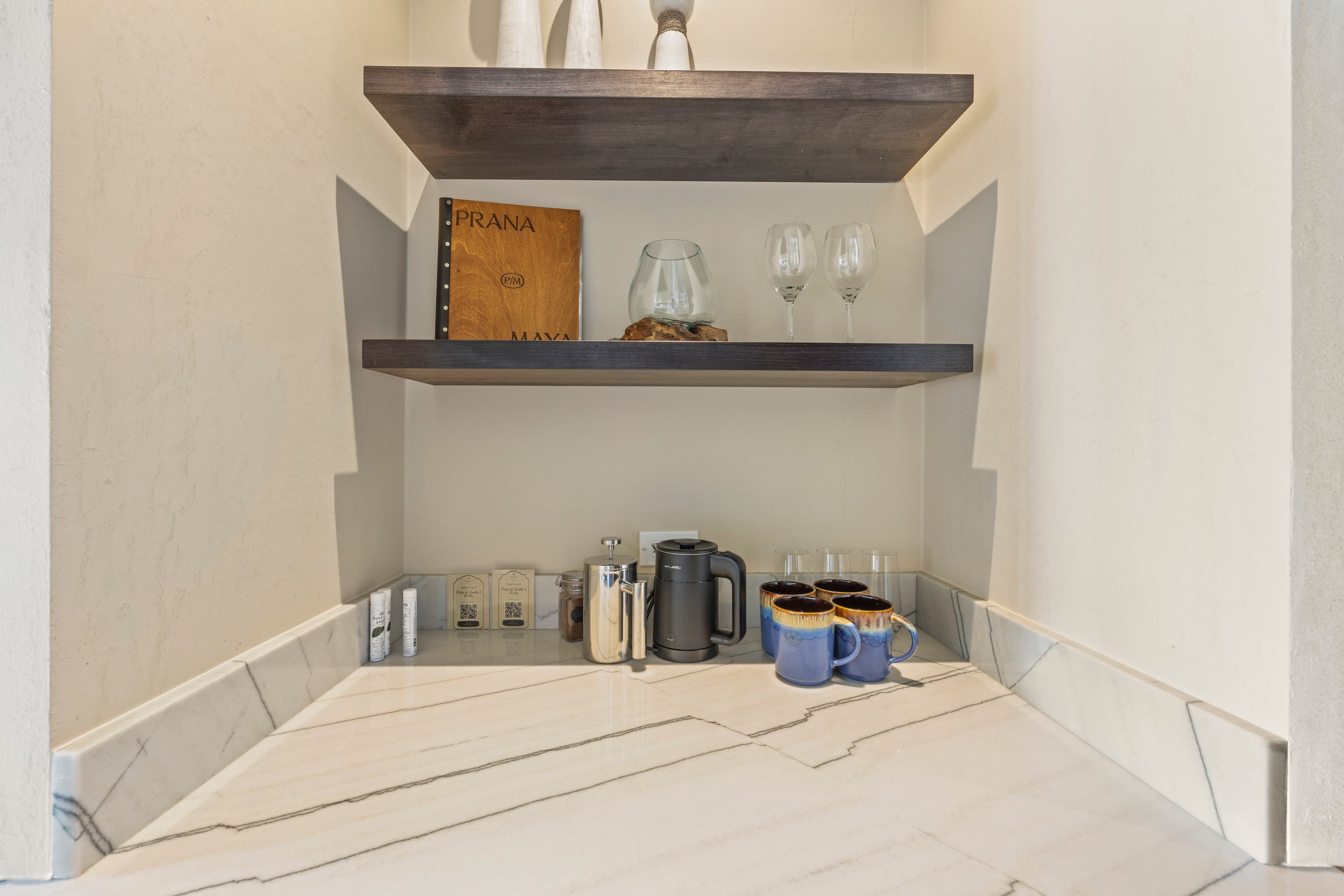 A cozy corner shelf featuring glassware, a book, and a coffee maker on a marble countertop.