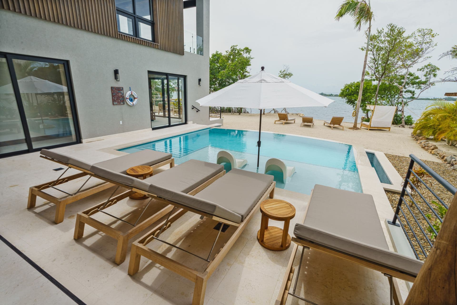 A poolside area features sun loungers, an umbrella, and views of greenery and water.