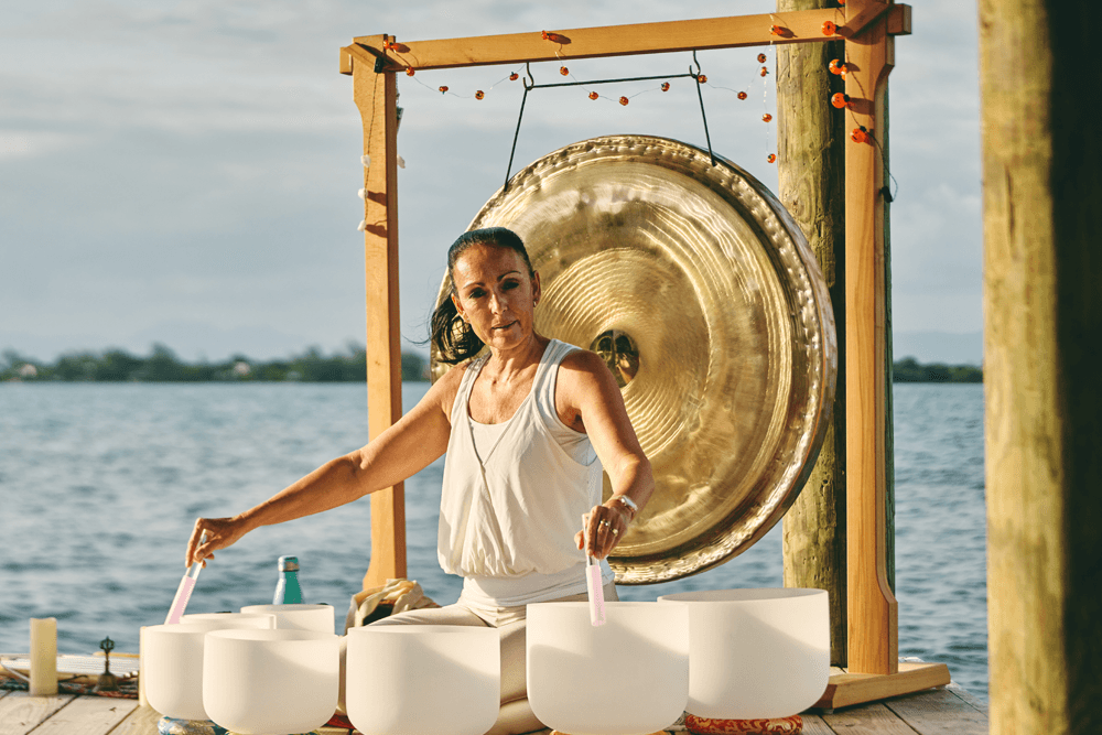 A woman plays crystal singing bowls and a gong by the water during a sound healing session.