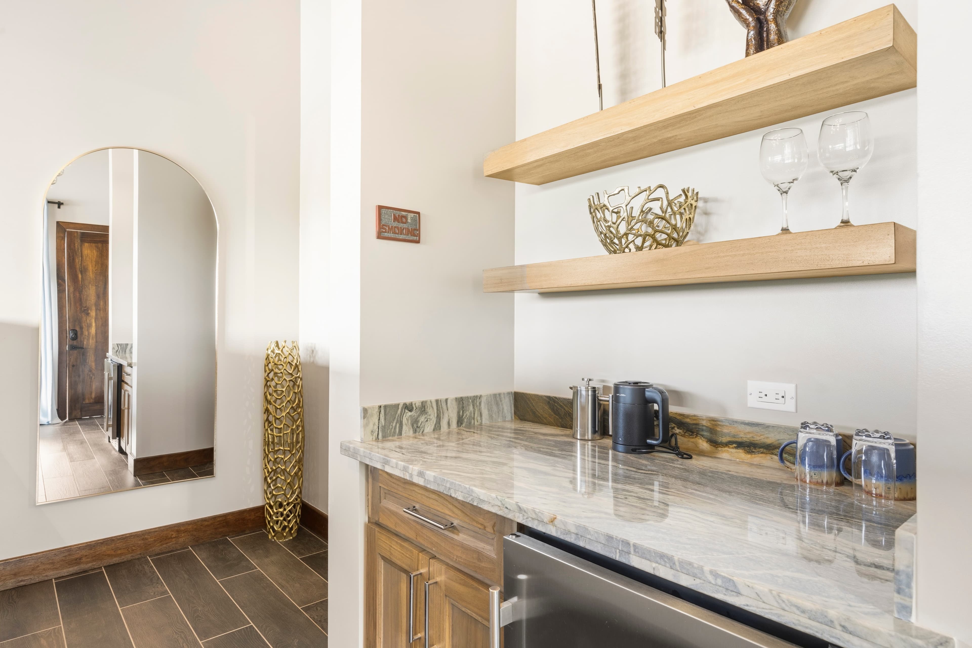 A modern kitchenette featuring wooden shelves with decorative items, glassware, and a sleek countertop.