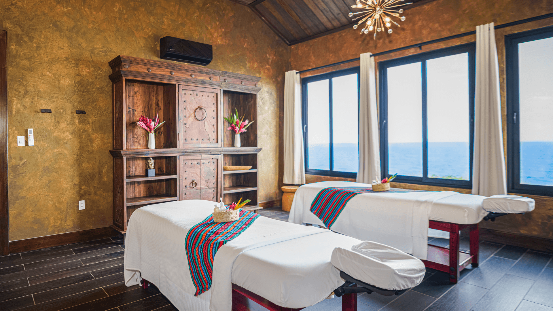 A serene spa room with two massage tables, tropical flowers, and ocean views.