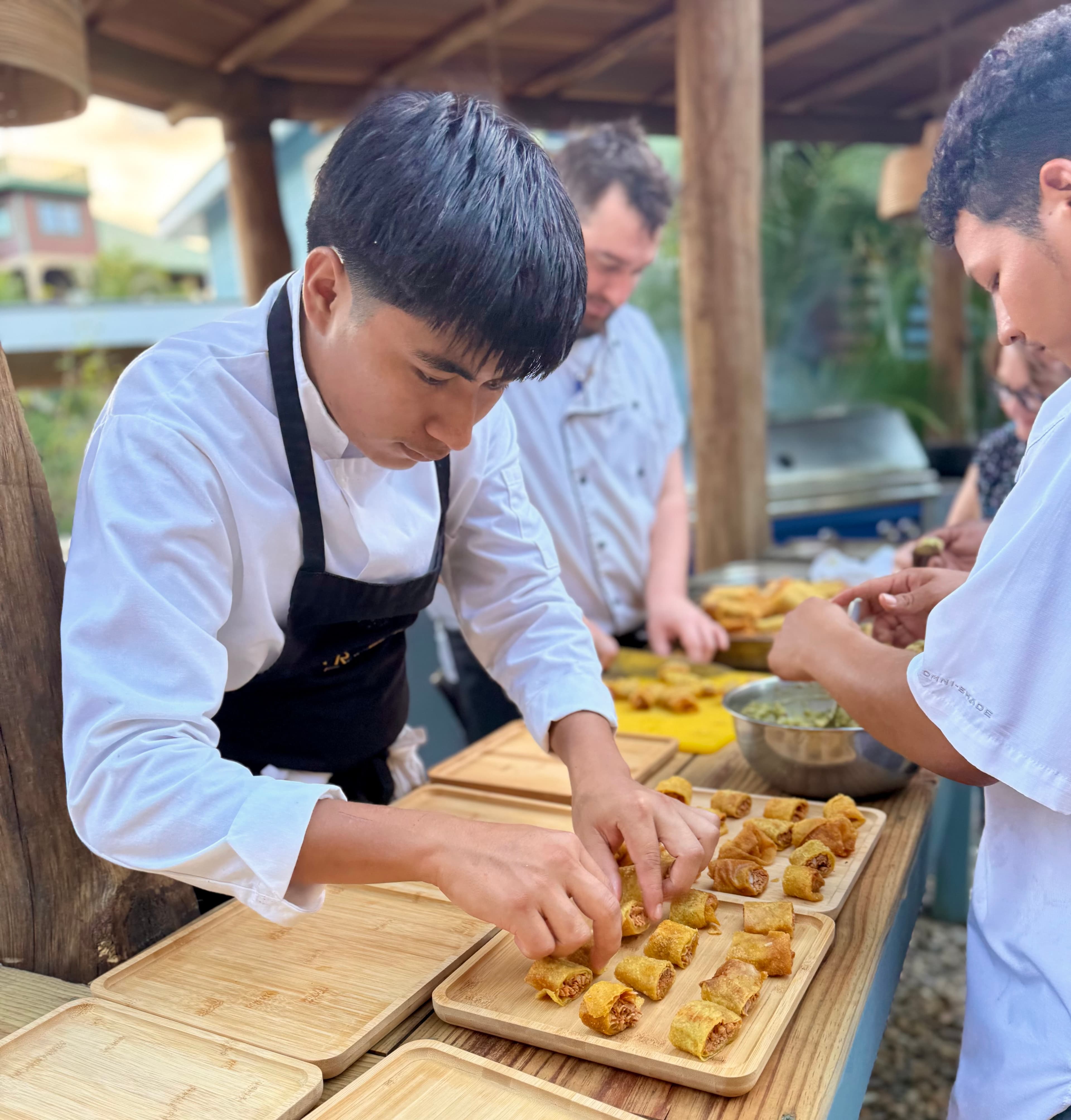 Chefs prepare spring rolls at an outdoor kitchen, focusing on meal preparation.