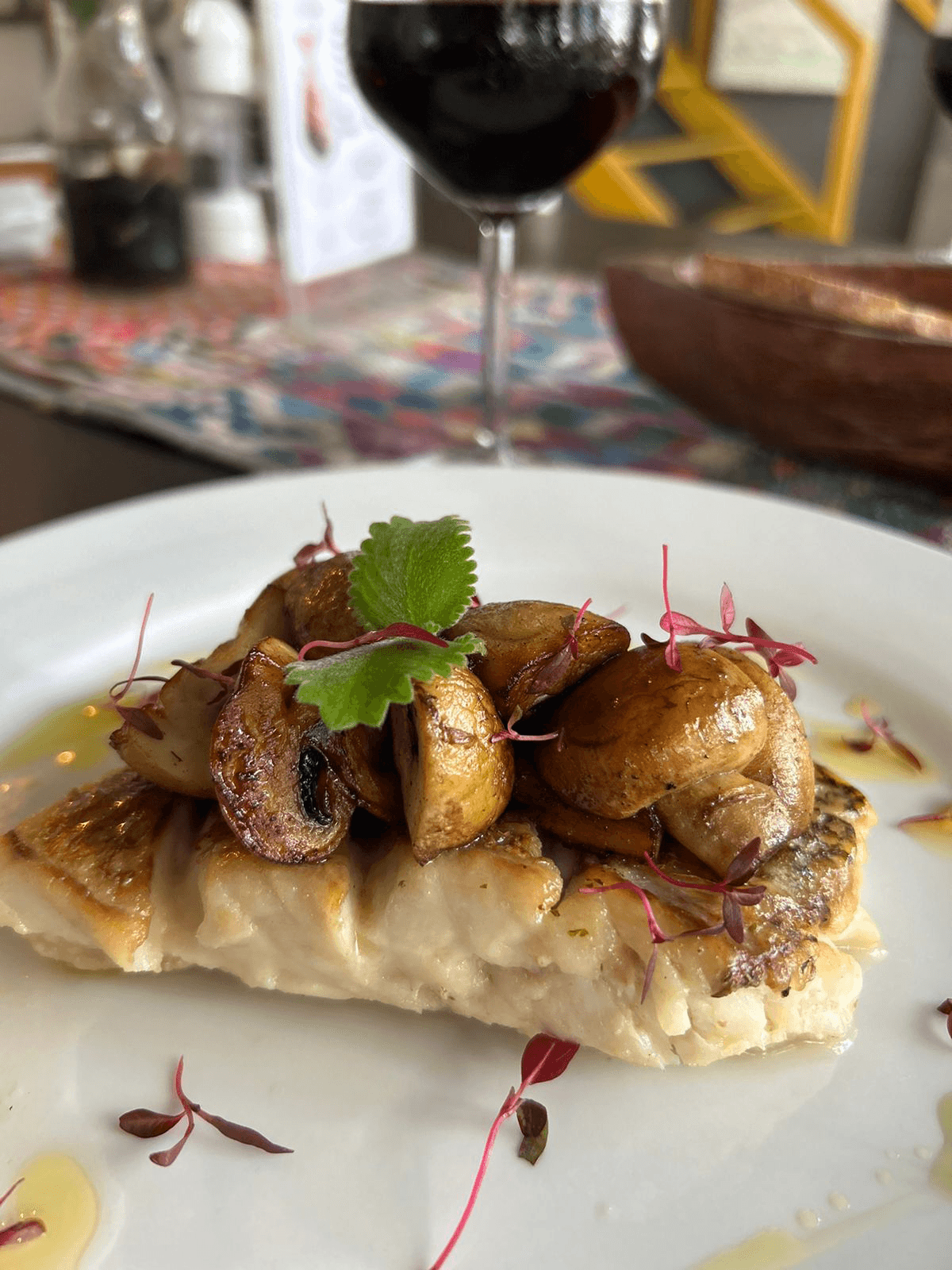 A plate of grilled fish topped with sautéed mushrooms and garnished with microgreens, accompanied by a glass of red wine.