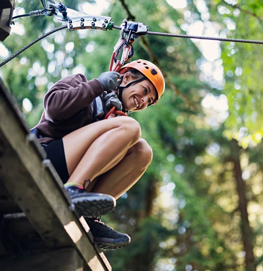 A smiling person in an orange helmet prepares to zip line through a forest.