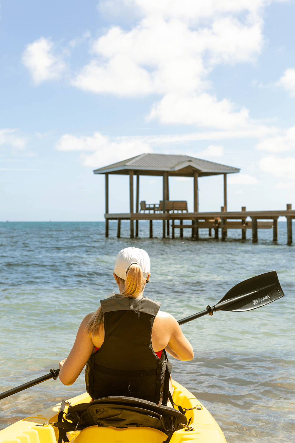 A person in a kayak paddles towards a wooden pier in clear blue water.