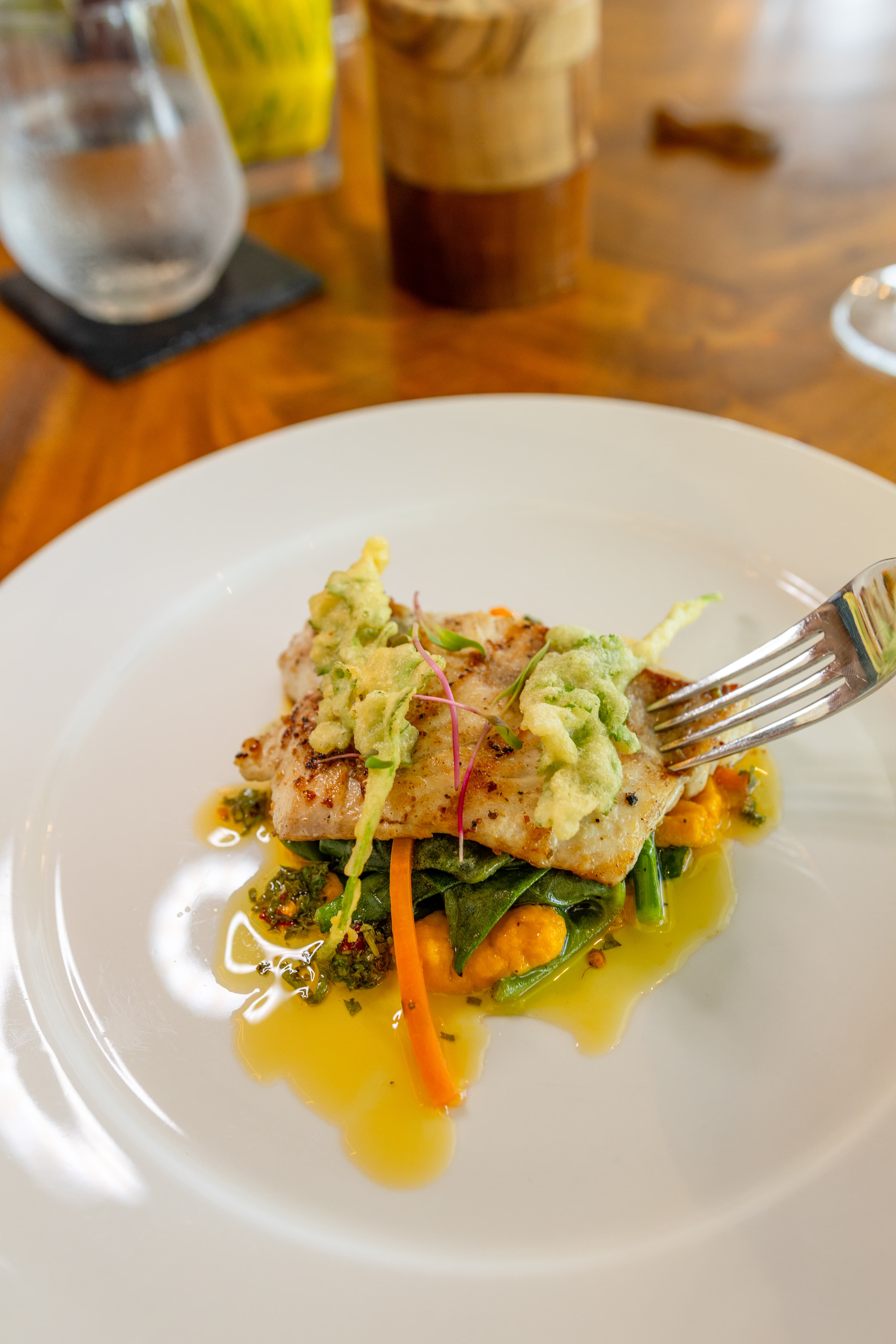 A plated fish dish topped with green sauce, accompanied by colorful vegetables and drizzled with olive oil.