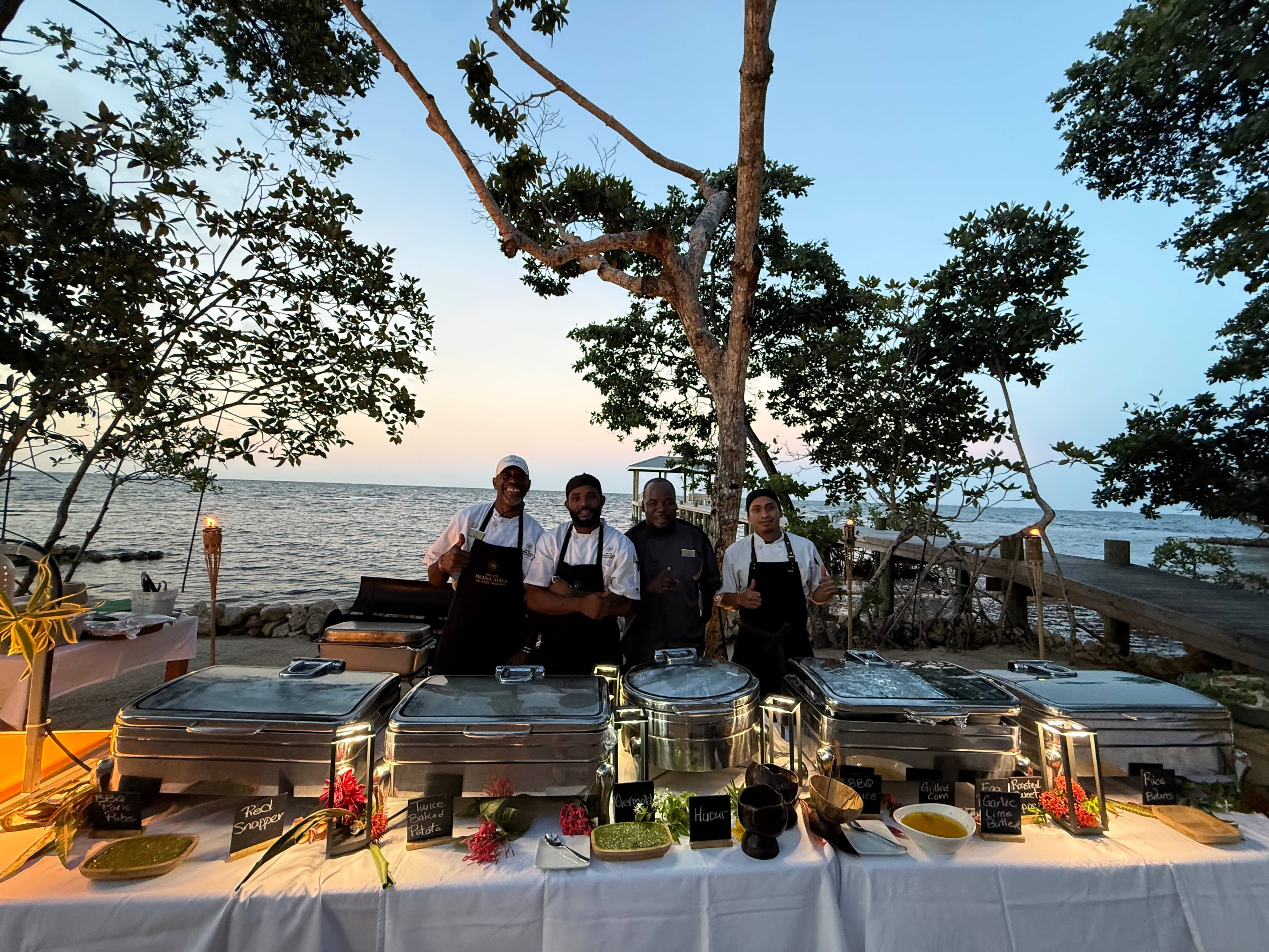 Three chefs stand behind a buffet setup by the beach at sunset.
