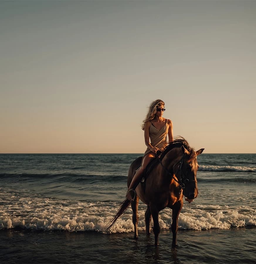 A woman in a beige outfit rides a horse in shallow waves during sunset on a beach.