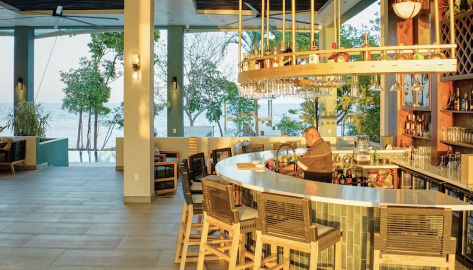 A bartender prepares drinks at a stylish bar with a view of the ocean and greenery.