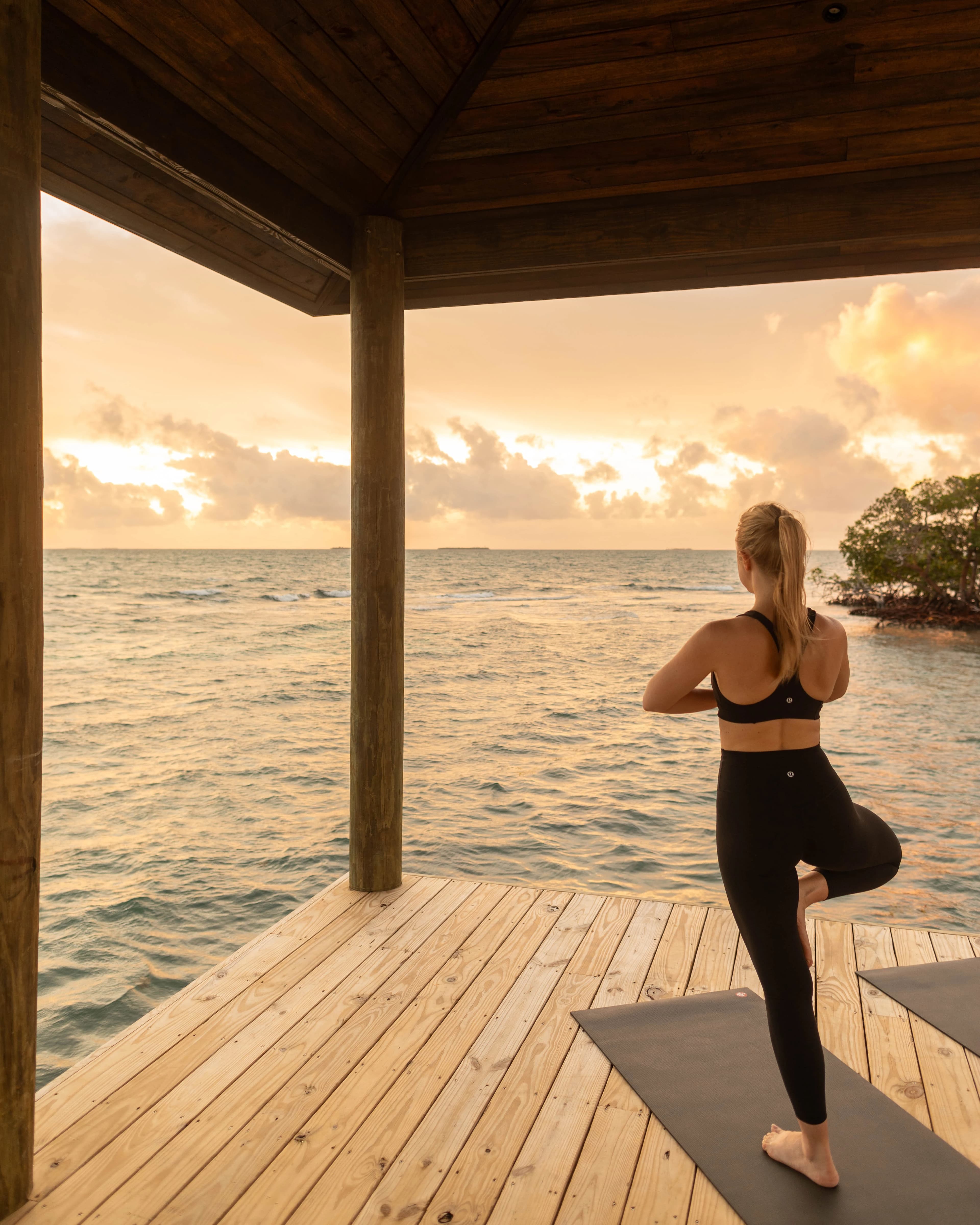 A woman practices yoga on a dock overlooking the ocean during sunset.