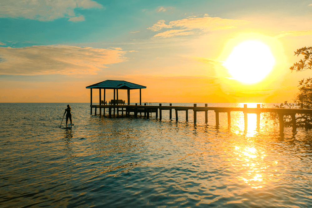 A person paddleboards near a wooden pier at sunset, with vibrant colors reflecting on the water.