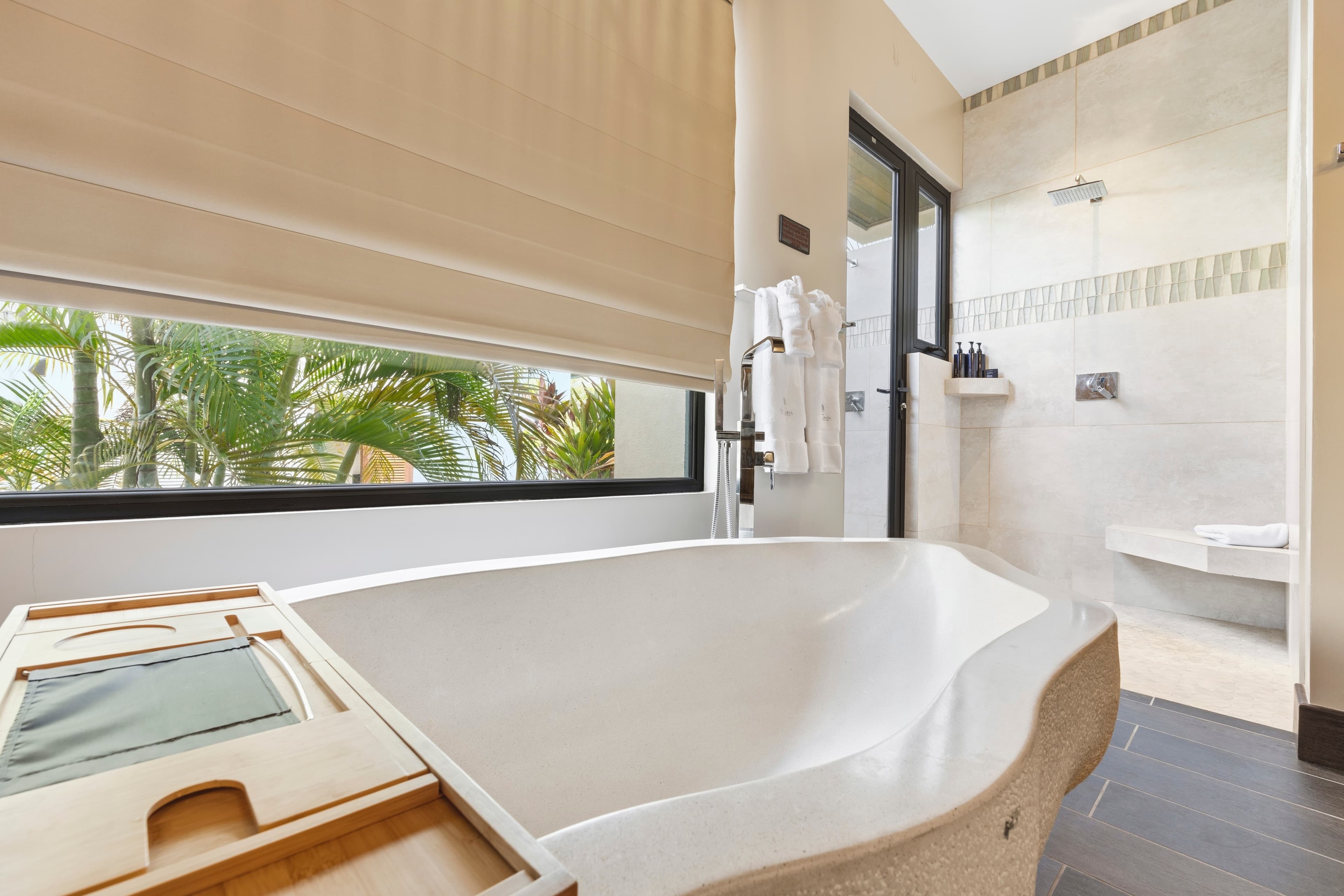 A modern bathroom featuring a freestanding tub, a glass-enclosed shower, and natural light from a window with palm trees visible outside.