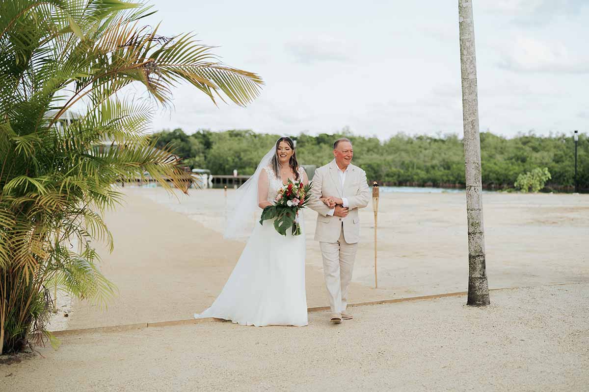 A bride in a white gown walks happily down an outdoor aisle with her father.