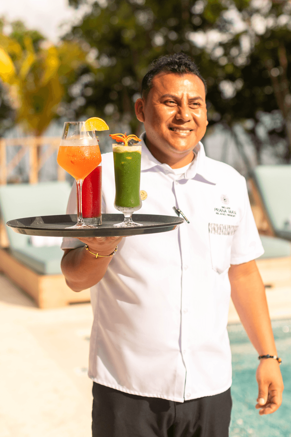 A smiling server holds a tray with a colorful cocktail and a green drink by a pool.