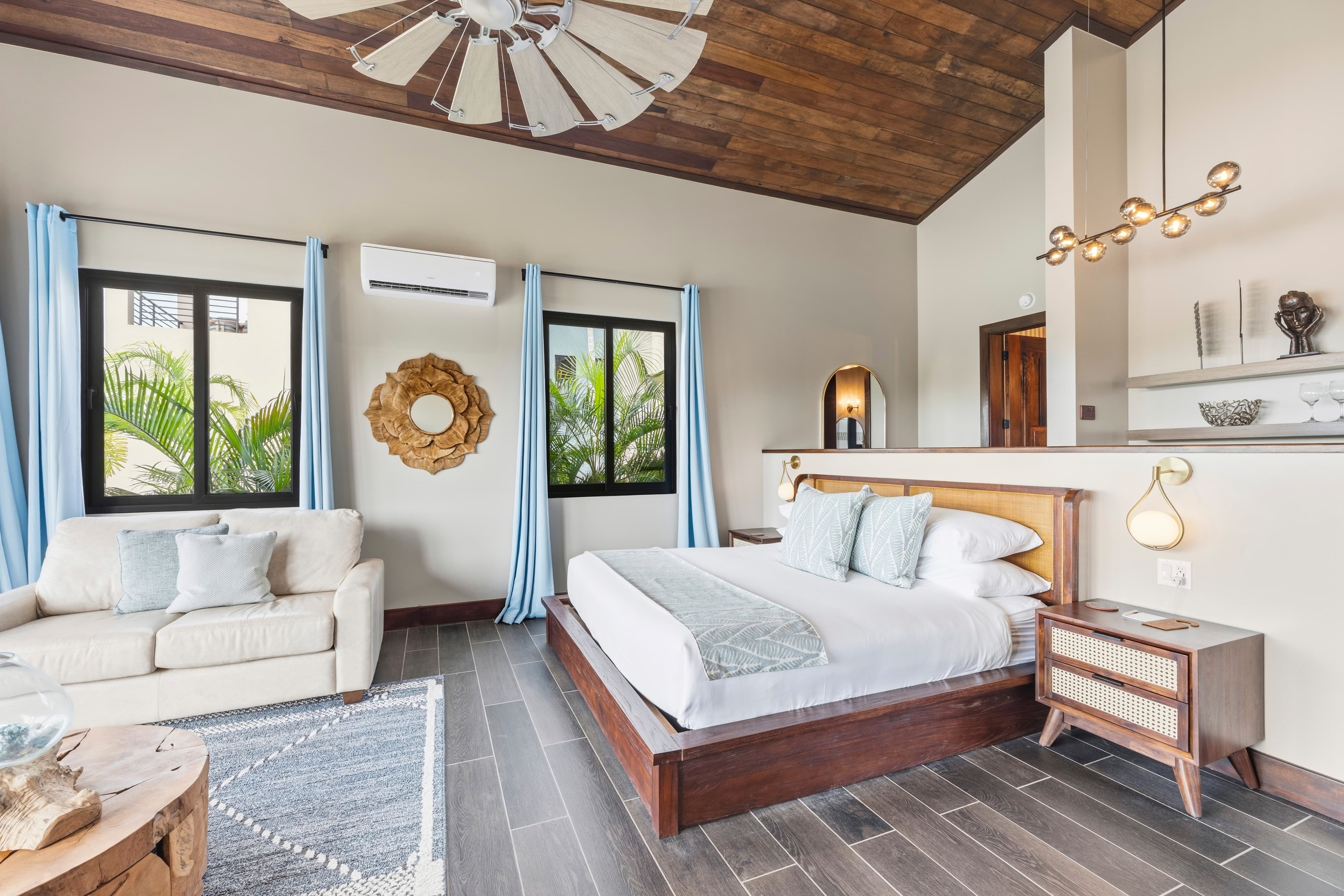 A spacious and modern bedroom featuring a bed, sofa, and large windows with light blue curtains.