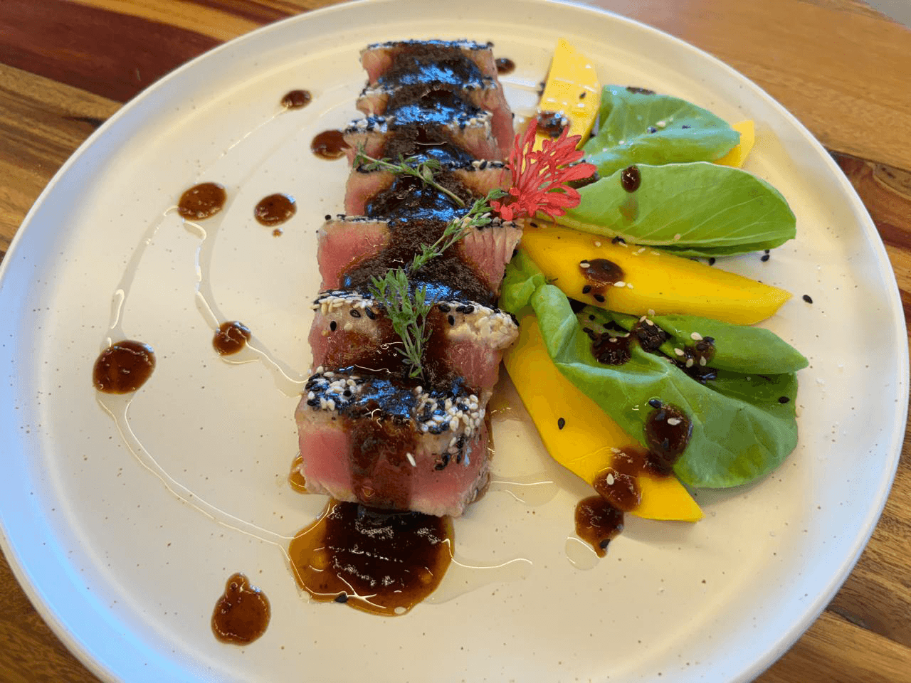A plate of seared tuna slices garnished with sesame seeds, mango slices, greens, and drizzled with sauce.