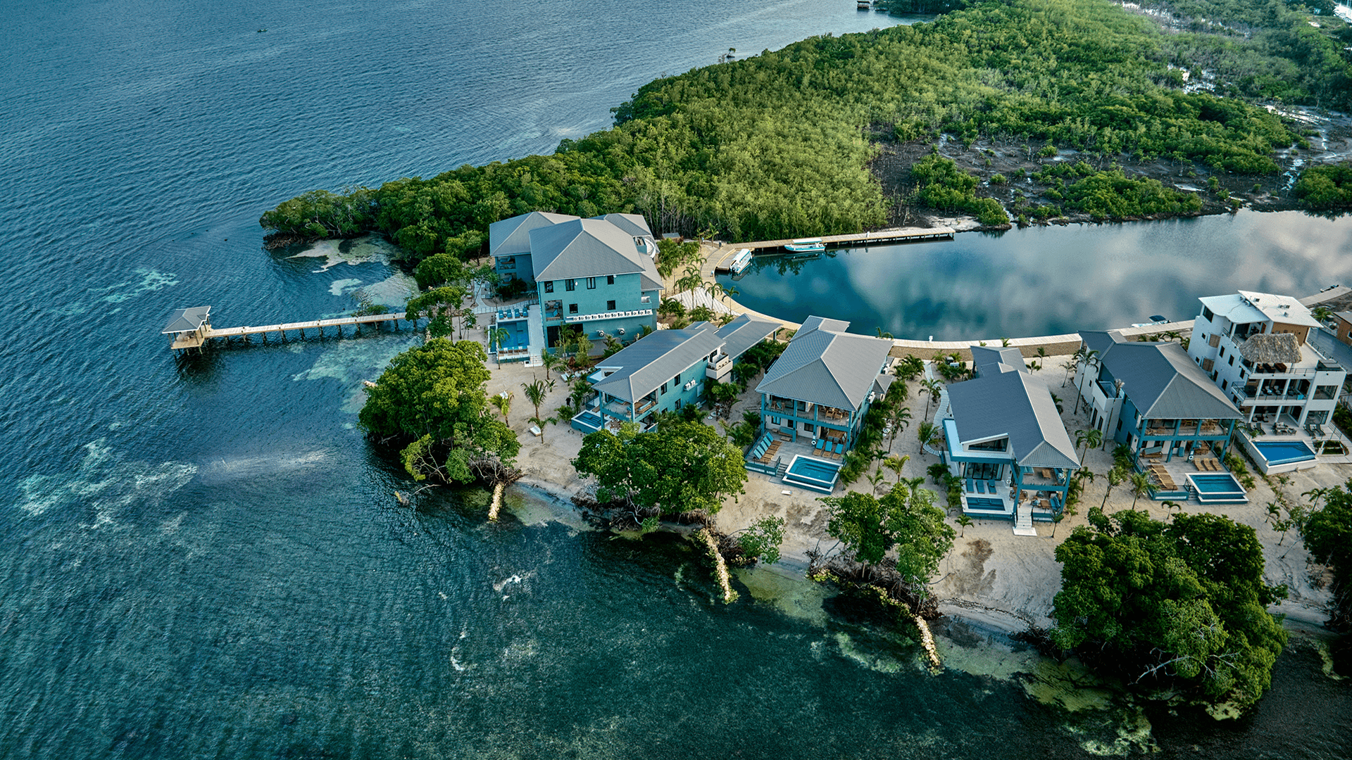 An aerial view of a tropical beachfront resort featuring several homes and a pier surrounded by water and lush greenery.