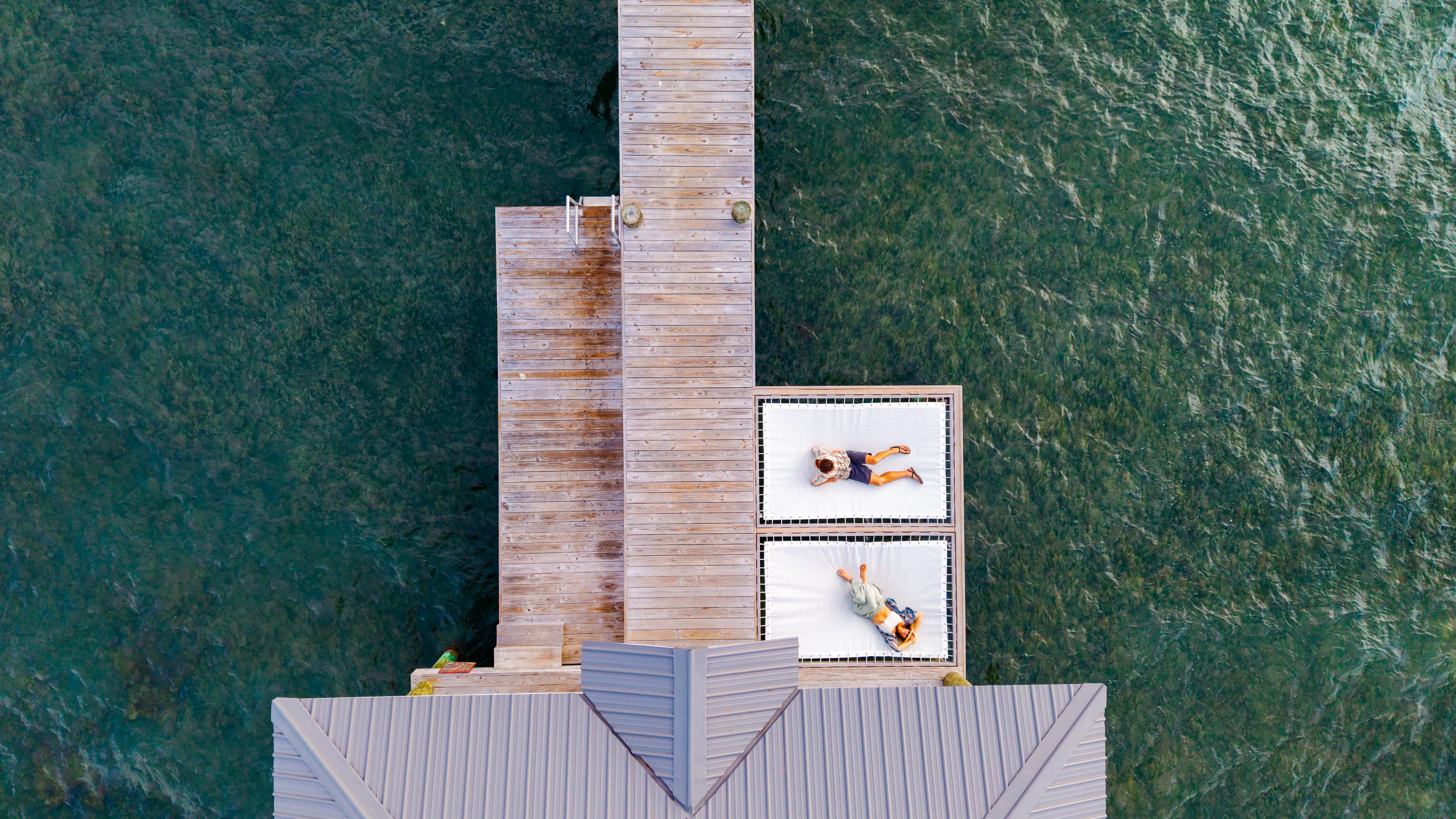 Aerial view of two people lounging on sunbeds next to a wooden dock over clear water.