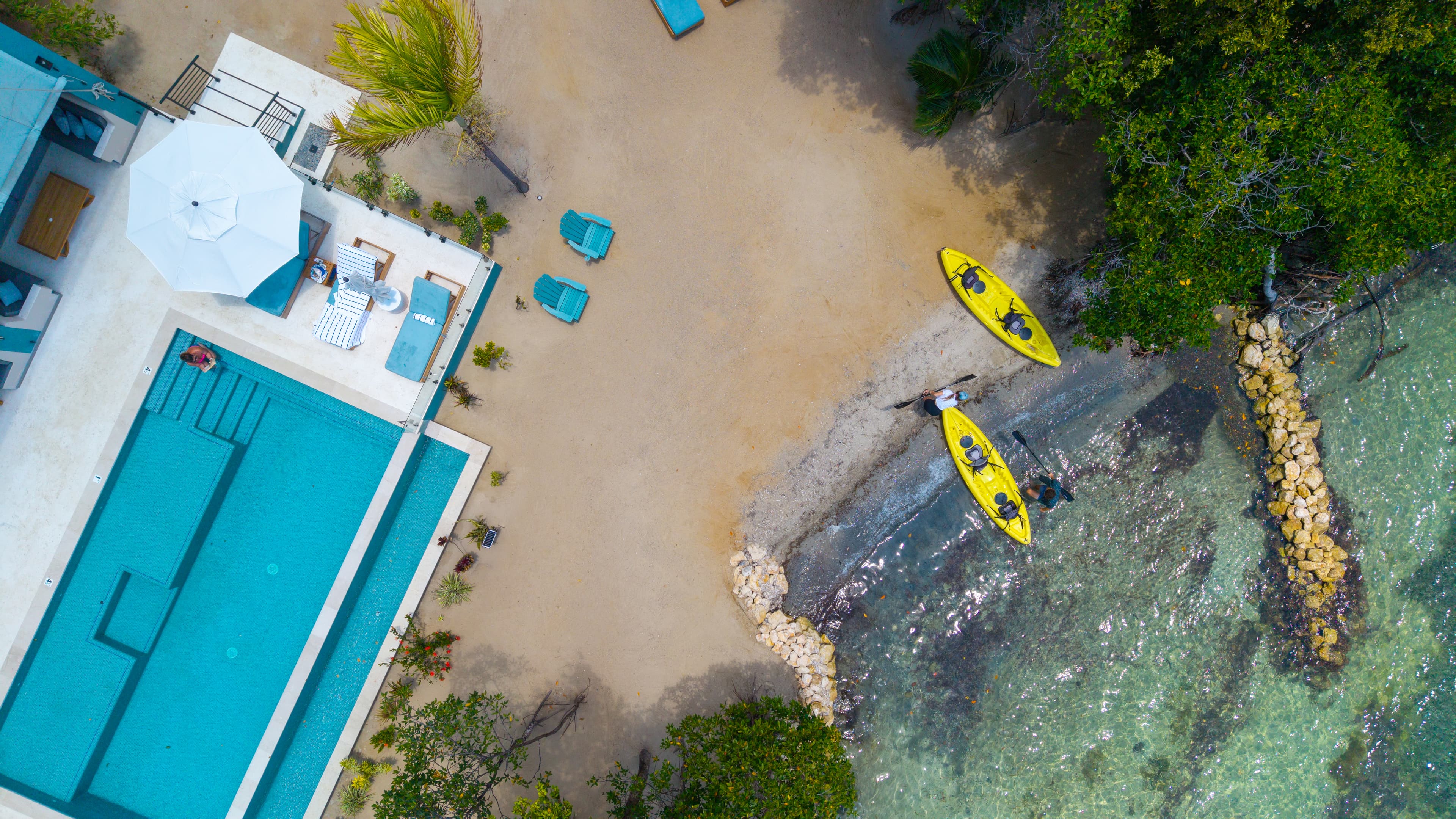 Aerial view of a sandy beach with two yellow kayaks, a pool, and lounge chairs beside lush greenery.
