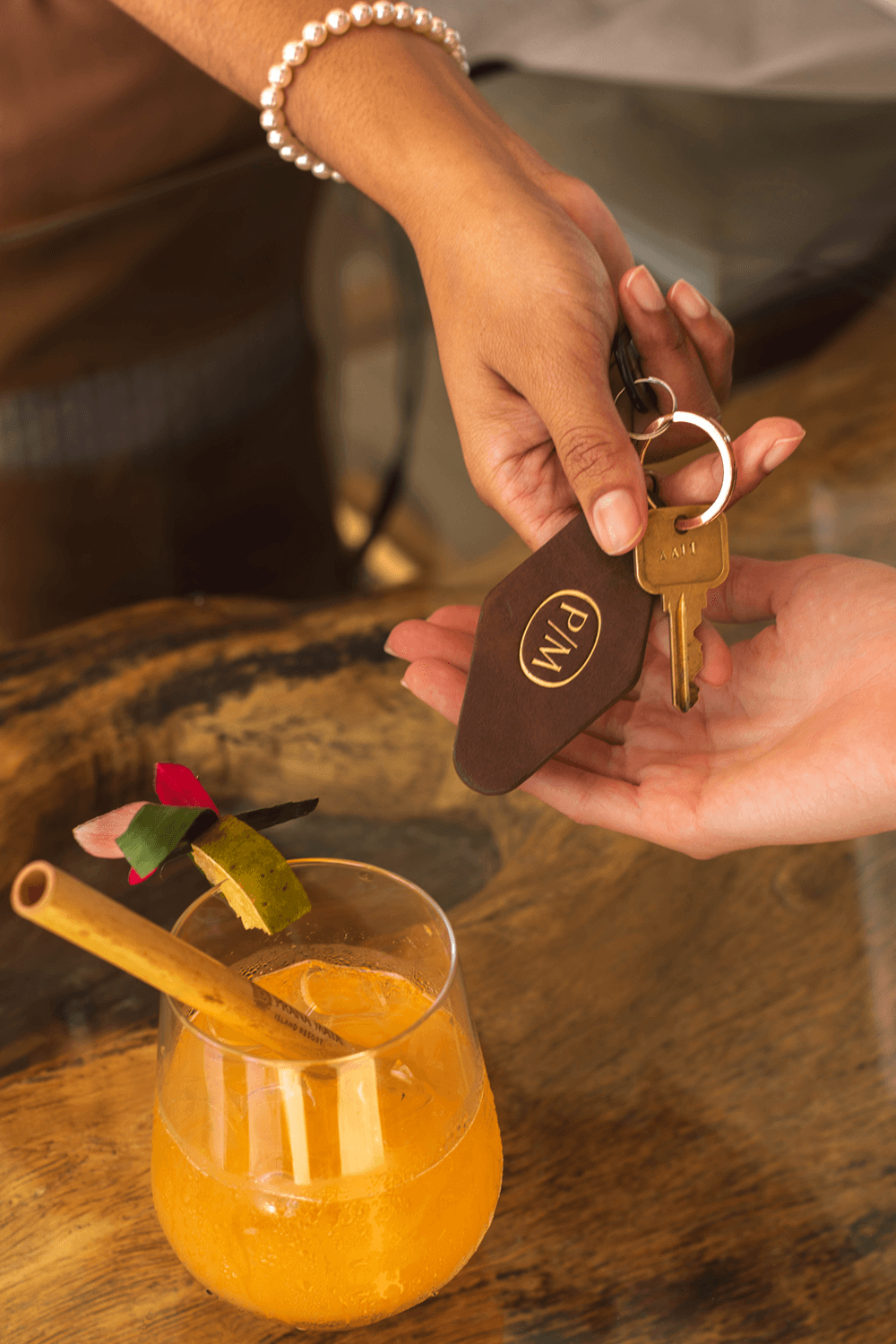 A hand is giving a leather keychain with the text "PIN" to another hand, with a tropical drink in the foreground.