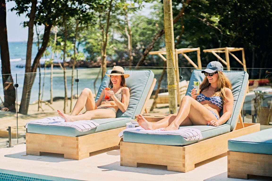 Two women relaxing on lounge chairs by the pool, each enjoying a colorful drink.