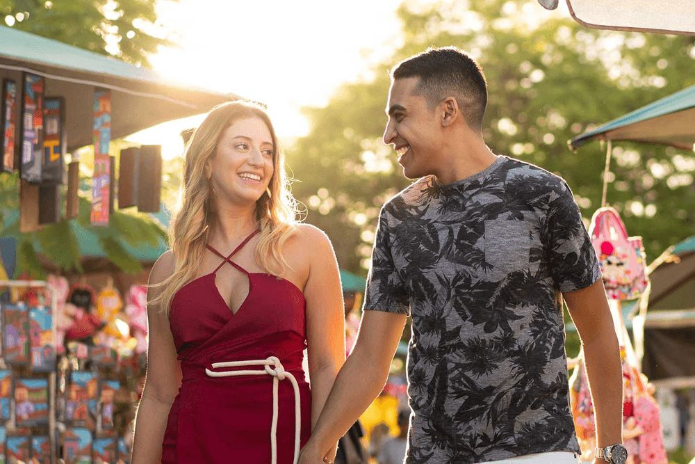 A couple walks hand-in-hand, smiling at each other in a vibrant outdoor market.