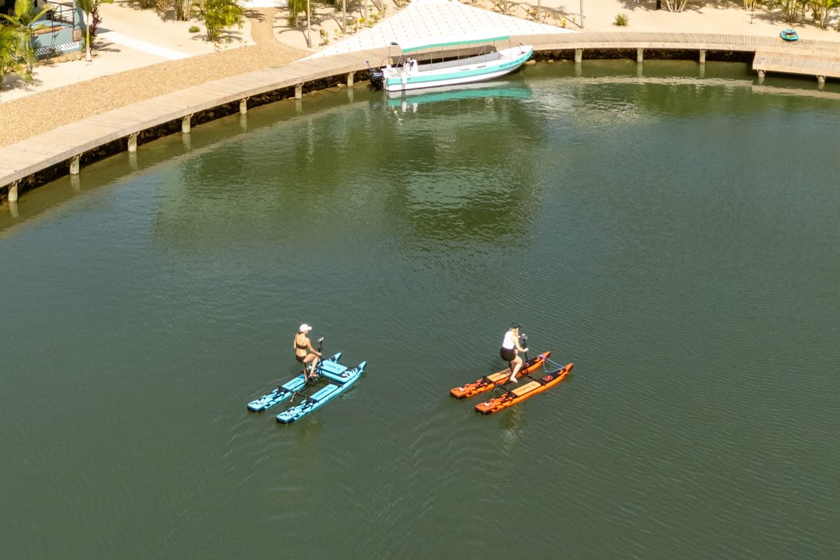 Two people paddle on bright blue and orange kayaks in a calm, greenish waterway.