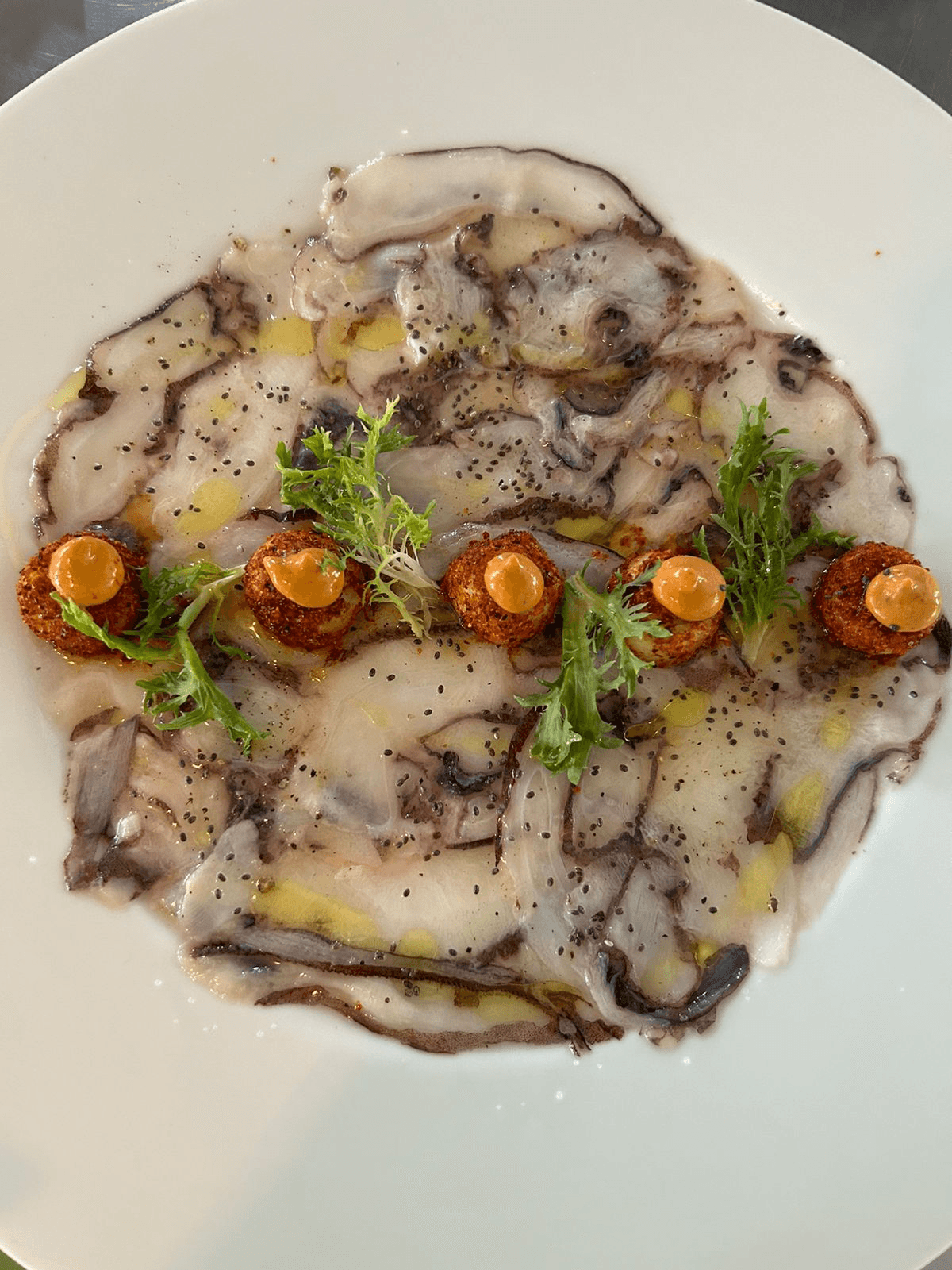 A plate featuring thinly sliced octopus carpaccio, drizzled with olive oil, topped with four seasoned round fritters and garnished with greens.
