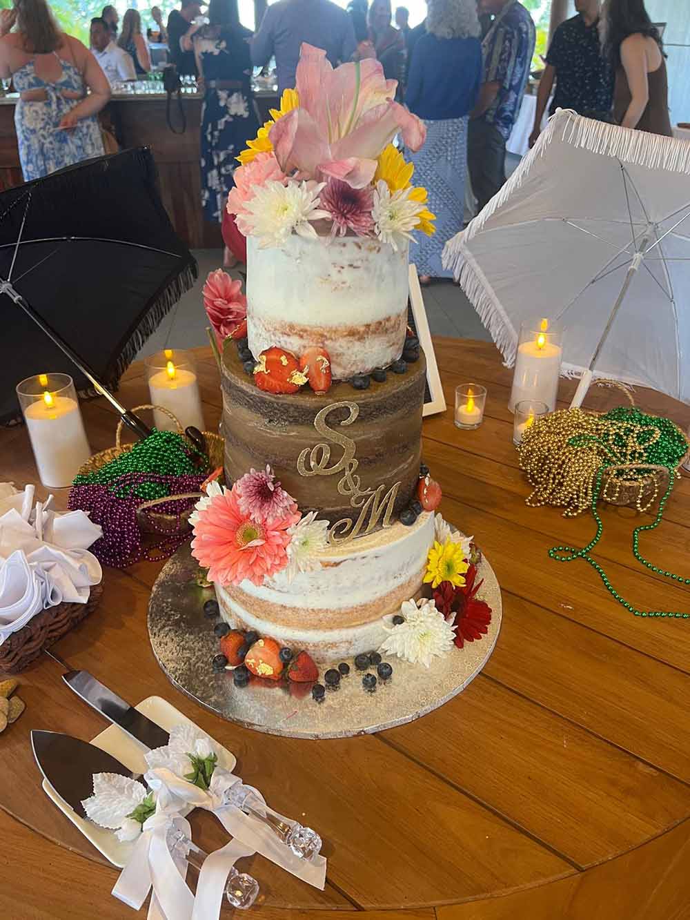 A three-tiered wedding cake adorned with flowers and strawberries, surrounded by decorative candles and beads on a wooden table.