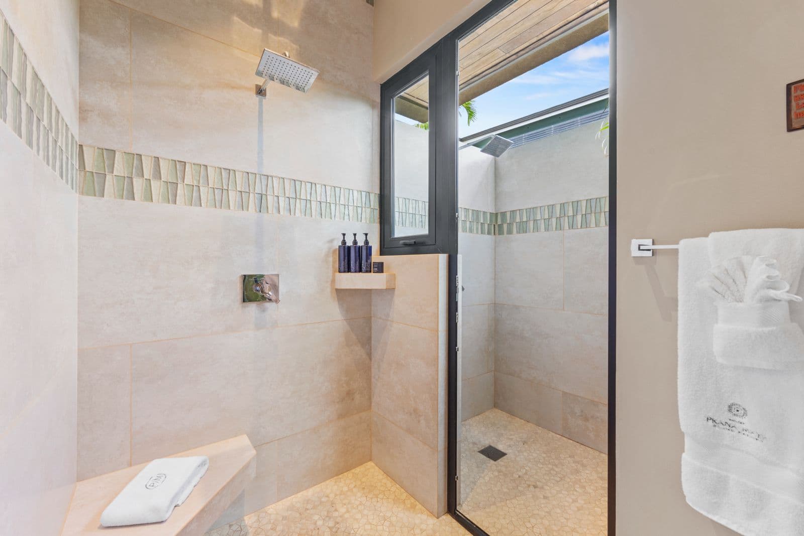 A modern, spacious shower with natural light, featuring stone walls, a rain showerhead, and neatly arranged toiletries.