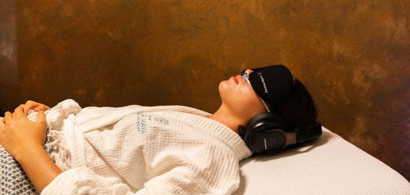 A person in a robe rests on a bed, wearing headphones and a sleep mask.