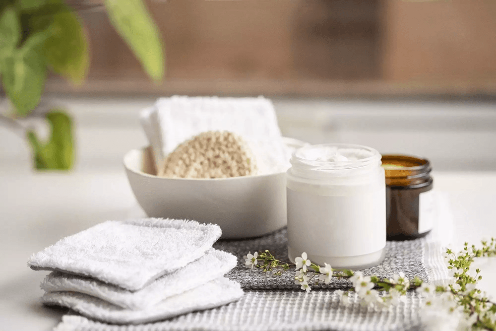 A collection of white towels, skincare products, and a small bowl with a natural sponge on a textured surface.