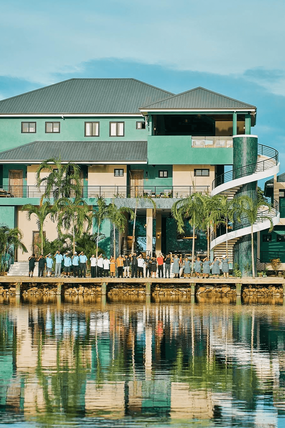 A group of people stands on a pier in front of a modern green building, reflecting in the water.