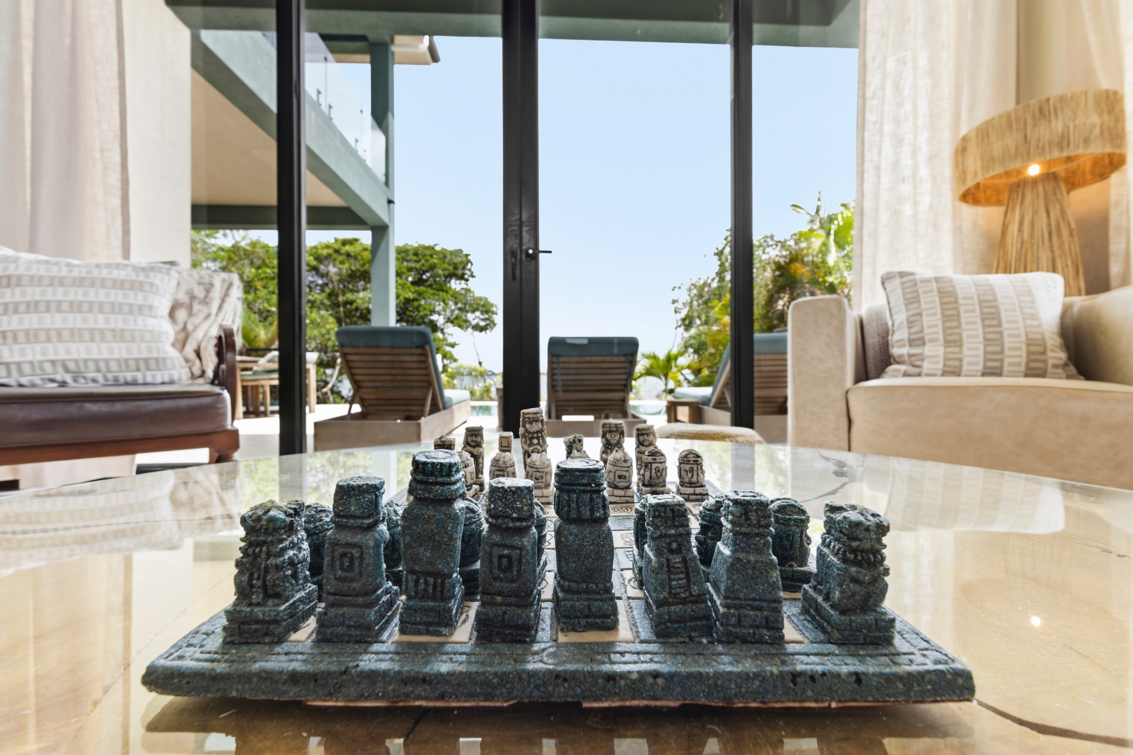 A unique chess set made of ornate pieces sits on a glass table in a bright, stylish living room.