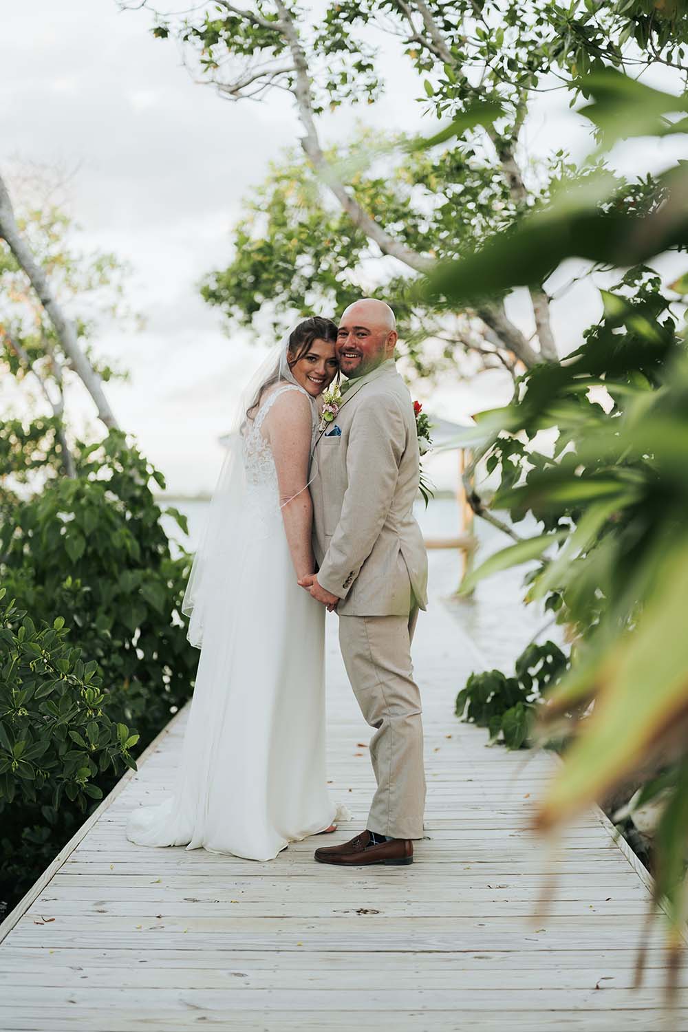 A happy bride and groom stand together on a wooden pathway surrounded by greenery.
