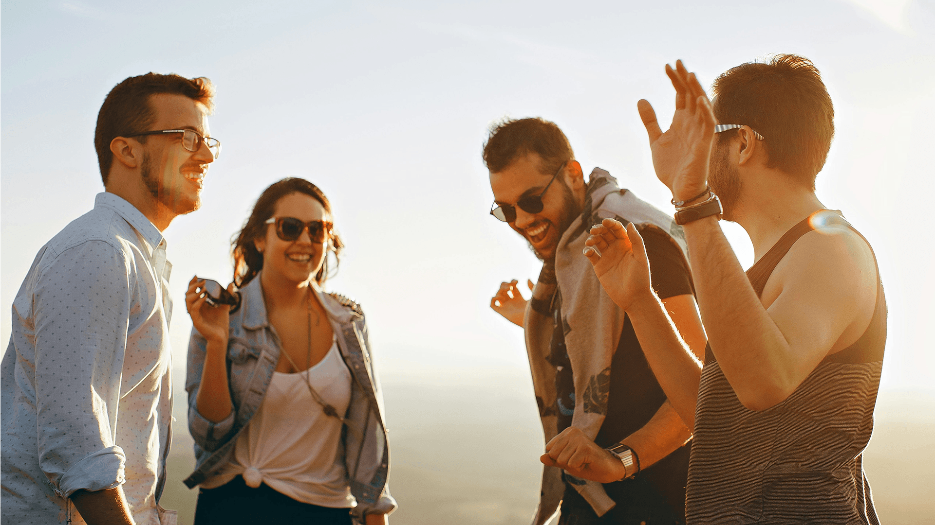 A group of four friends smiles and dances together outdoors against a sunset backdrop.