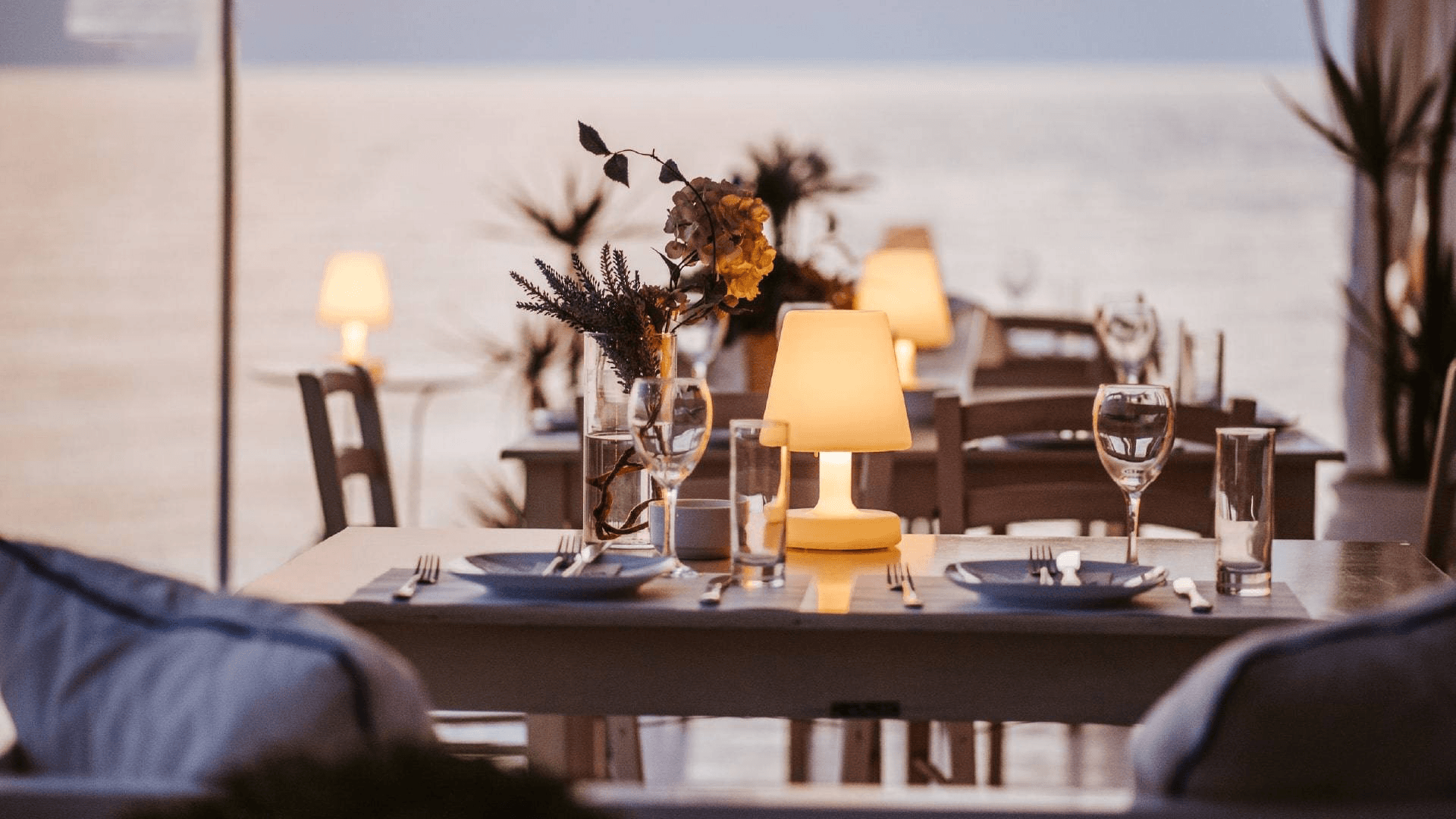 A romantic restaurant table set with soft lighting and flowers, overlooking a serene sea at dusk.