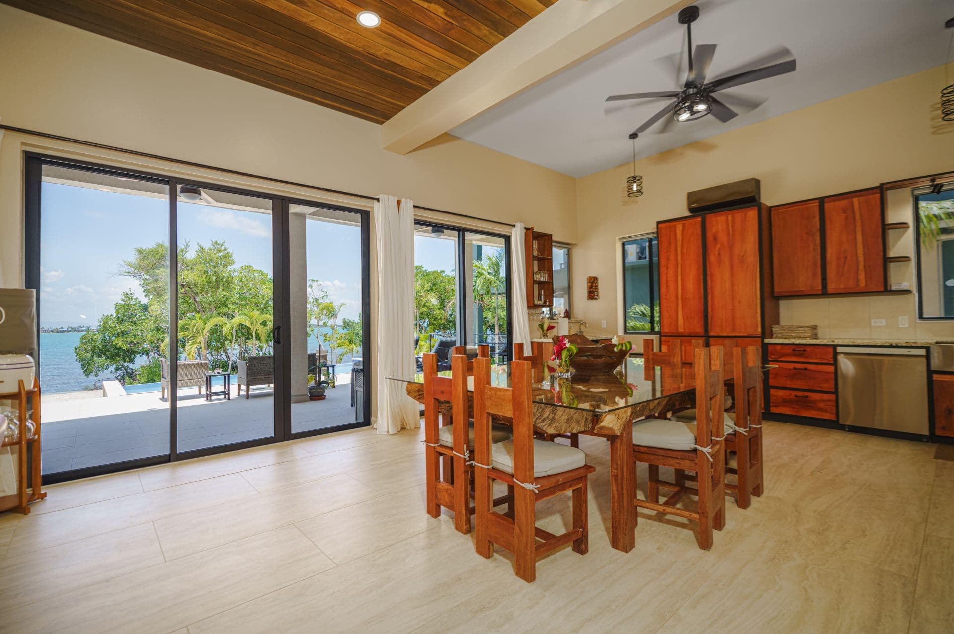 A spacious dining area featuring wooden furniture with a view of a tropical landscape through large sliding glass doors.
