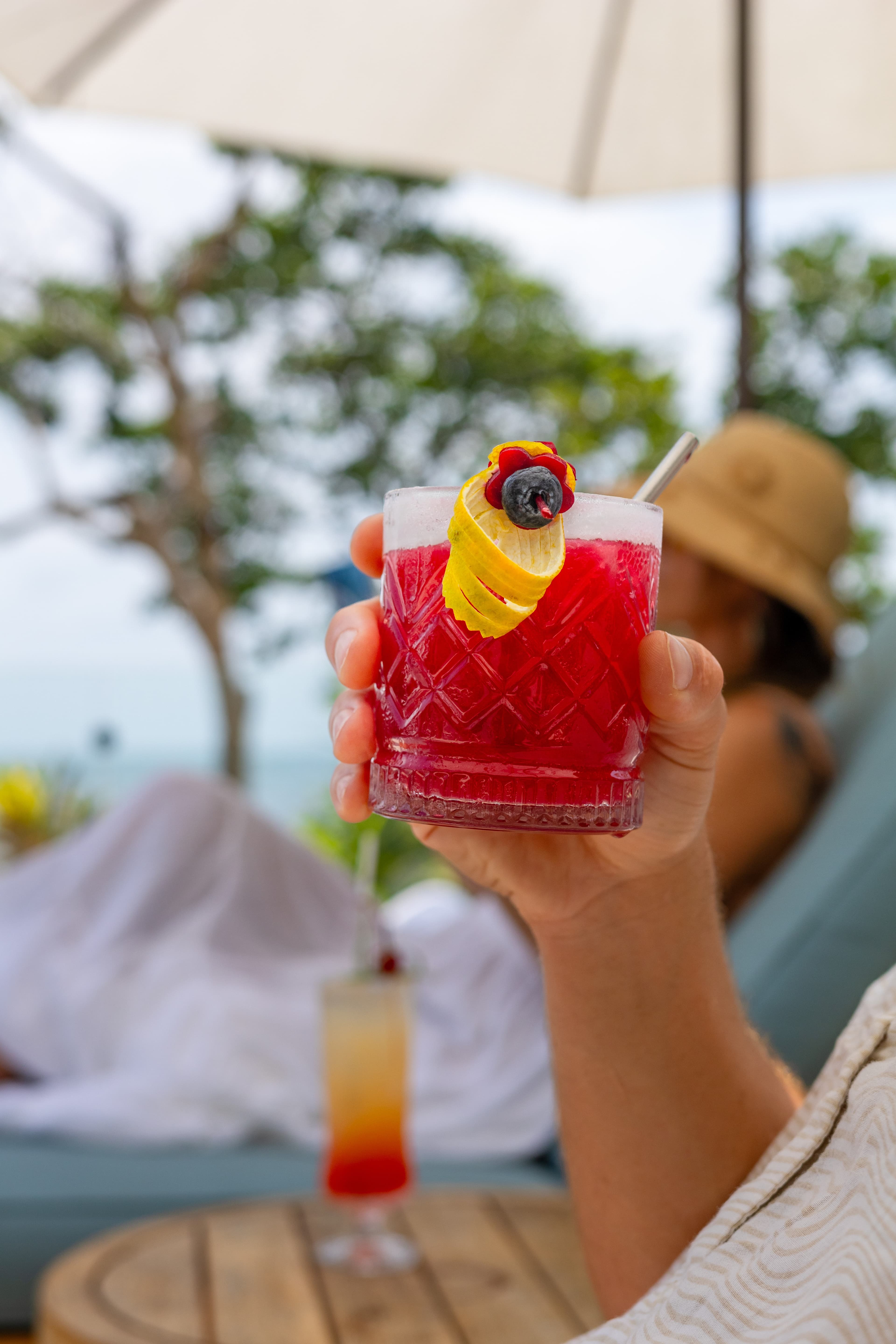 A hand holds a vibrant red cocktail garnished with a lemon twist and blueberry, with a blurred background of a tropical setting.
